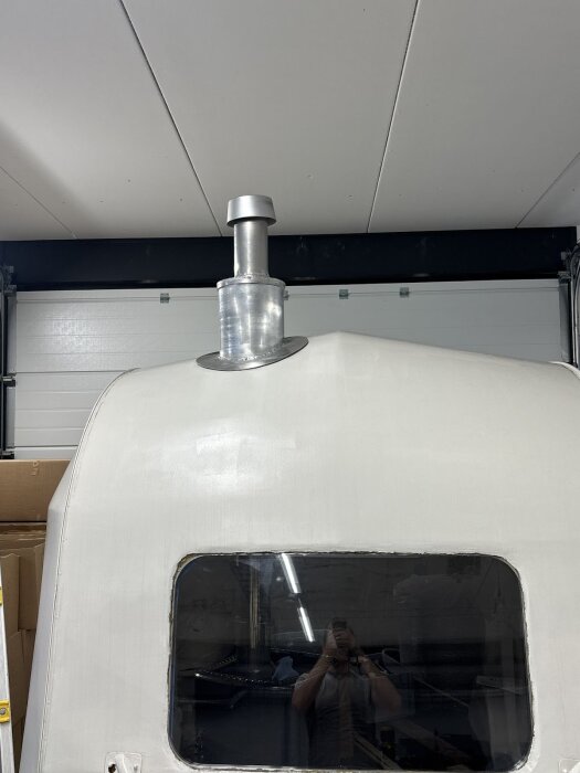 En monterad regnhuv av en kastrull på en skorsten ovanpå en vit yta, troligen en husvagn eller liknande. Vitt tak och fönster i bakgrunden.