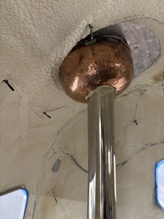 Rostfritt rökrör monterat i tak med kopparfärgad regnhuv och fixeringselement synliga, del av skorstenprojekt i inomhusmiljö.