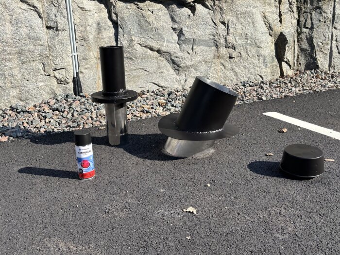 Svartmålat skorstensrör, värmefärg och kåpa ligger på marken vid en stenmur, del av ett byggprojekt.