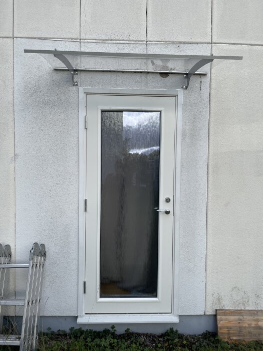 Vit dörr med glas i mitten sitter på en betongvägg. En liten skärmtak ovanför dörren. En hopfällbar stege står lutad mot väggen.