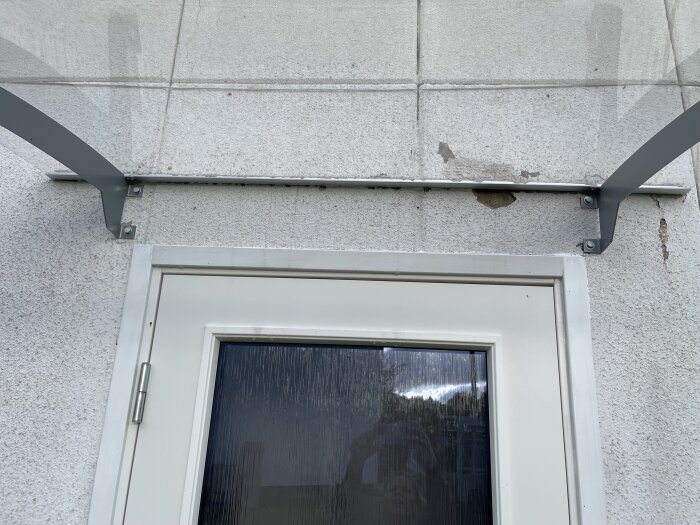 Överliggande baldakin med metallfästen ovanför en ytterdörr med laminerat glas, med skadad puts på väggen.