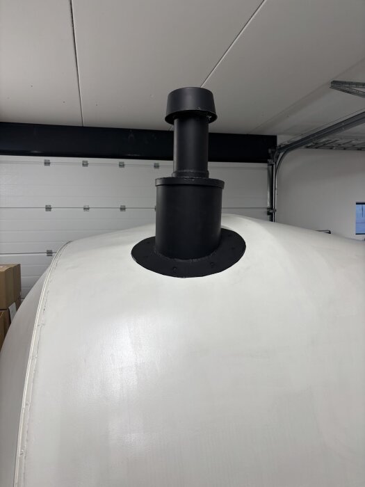 Svartmålad skorsten med regnhuv monterad på vitt tak inomhus; garage med vita väggar och tak.