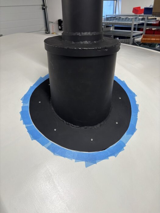 Svartmålad skorsten med krage monterad på taket, omgiven av blå maskeringstejp.