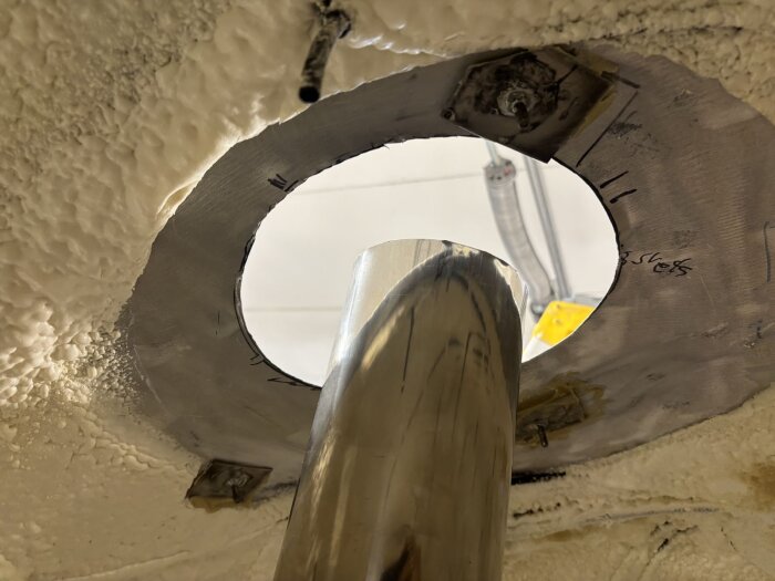 Rostfritt stålrör monterat genom isolerad öppning i taket, med metallbeslag och skruvar runt rörets kant.