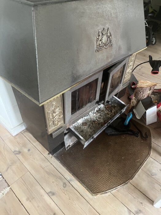 Gammal kamin med utsmyckat hölje och öppen lucka, placerad på trägolv med sotig insida synlig. Vedträn och verktyg ligger bredvid.