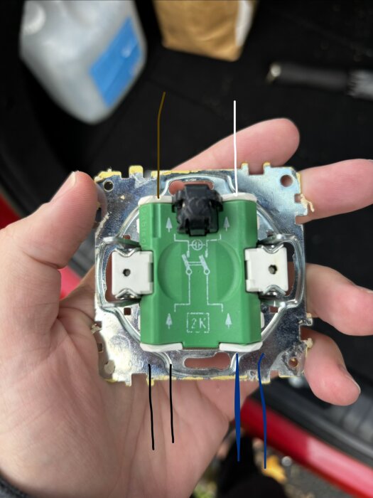 Närbild på en hand som håller en grön elektrisk komponent med olika färgade markeringar för ledningar: brun, vit, svart och blå.