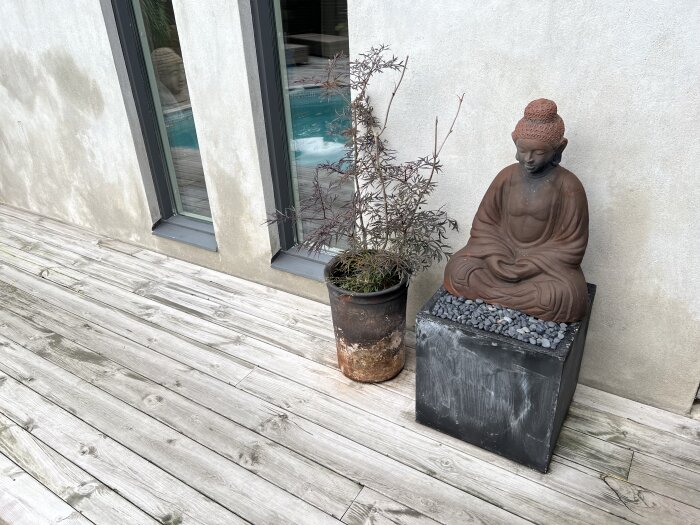 Putsad vägg vid trädäck med fuktmärken, bredvid fönster finns en Buddhastaty i brunt och en kruka med planta.
