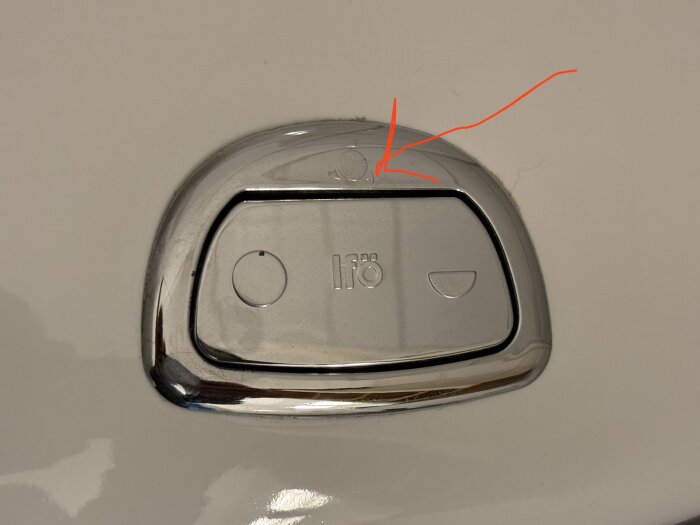 Kromad knapp med debossade symboler och markerat litet märke överst, pil visar platsen.