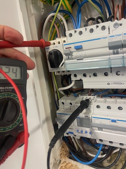 Multimeter mäter 225 VAC i elcentral. Två mätkablar är anslutna till fas och blå sladd på Astro-uret. Bakgrund av kablar och brytare.