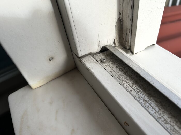 Fönsterkarm med tecken på vattenskada, inklusive sprickor och missfärgning på träet, nära ett hörn av ett öppningsbart fönster.