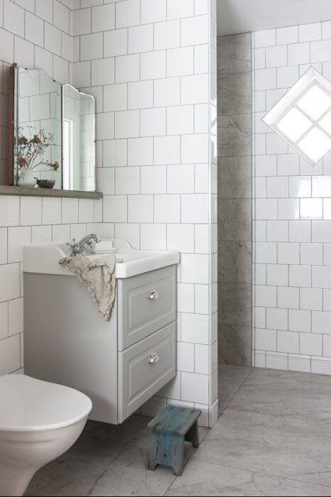 Badrum med vitkaklade väggar, handfat, spegel och grått skåp. Dusch med stenliknande kakel och ett litet fönster. Grått klinkergolv syns.