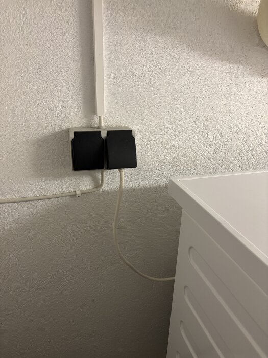 Vägguttag vid plats för tvättmaskin med synlig elinstallation och vit sladd som löper från ett av uttagen.