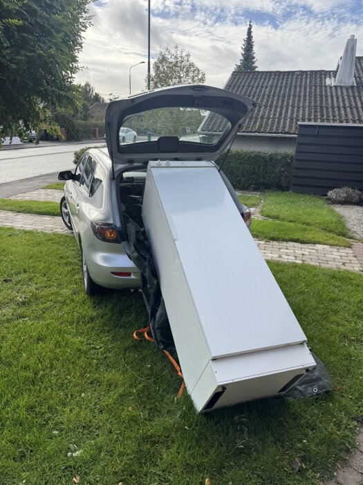 Torkskåp lastas ut ur en bil, bakluckan är öppen och torkskåpet lutar mot gräset i trädgården.