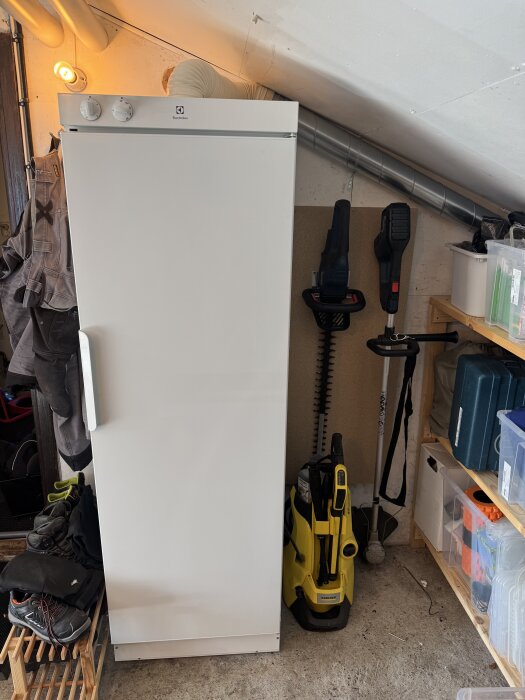 Vit Electrolux torkskåp i ett garage med verktyg, kläder och hyllor runtom, monterat med ventilationsrör i taket.