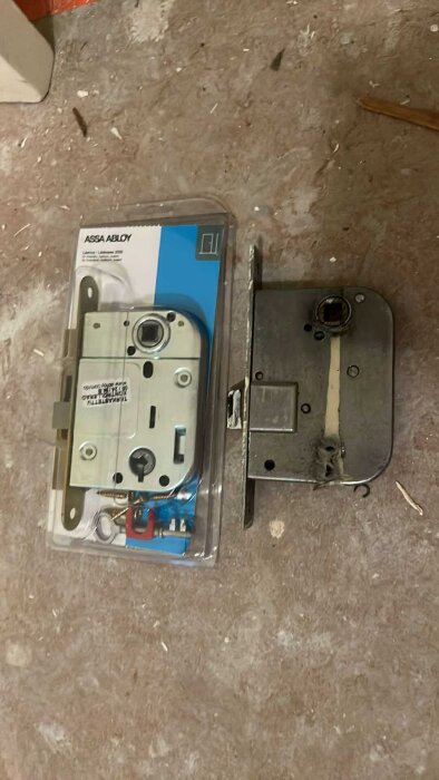 Två dörrlås visas, ett i förpackning och ett gammalt bredvid, på ett dammigt golv.