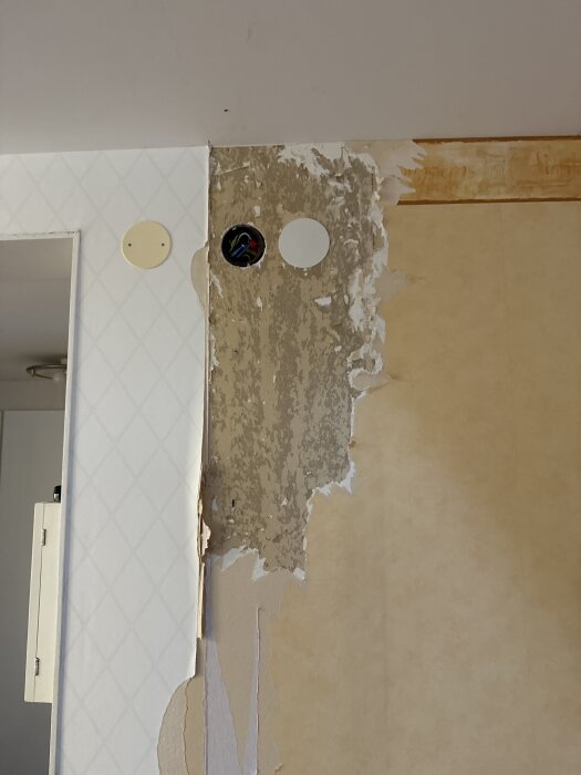 Exponerad vägg med borttagen tapet, gipsskivor synliga och två runda eluttag på vänster sida. Tapet i lager syns på höger sida.