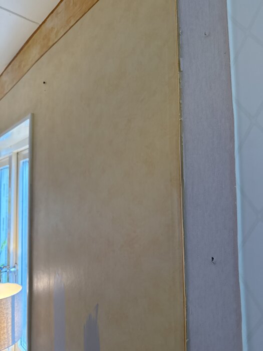 Vägg med tre lager tapet på gipsskiva, delvis målad med beige och blått, i ett rum med fönster och lampa, under renovering.