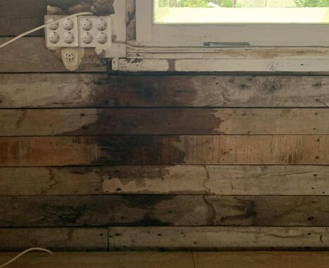 Vattenskada vid fönstret med mörka fläckar på träpanel under, troligtvis fuktskada från läckage. Flera eluttag synliga ovanför skadan.