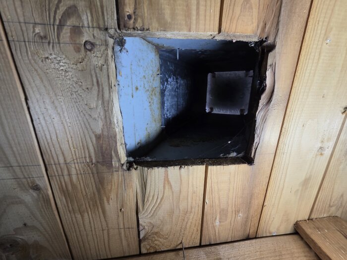 Ventilationsutlopp med rektangulär form på insidan av ett trätak, delvis skadat runt kanterna, för anslutning till köksfläkt.