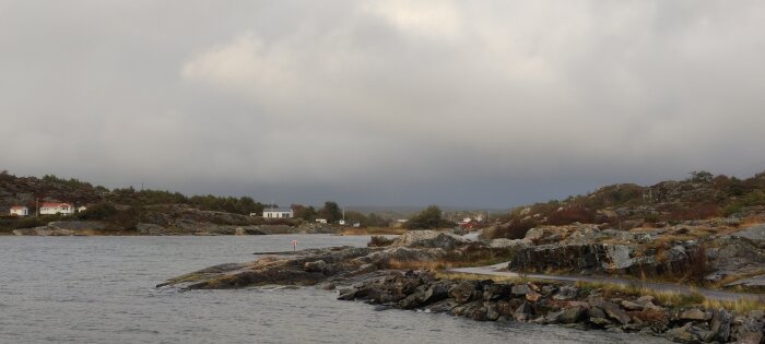 Gråmulen himmel över ett klippigt kustlandskap med hus, sett från vattnet.