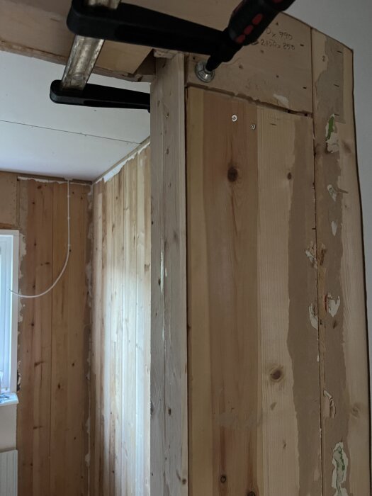 Trädörrkarm under byggnation med skruvade och limmade reglar, fastklämd av ett tving för stadga, omgiven av träpanelväggar.