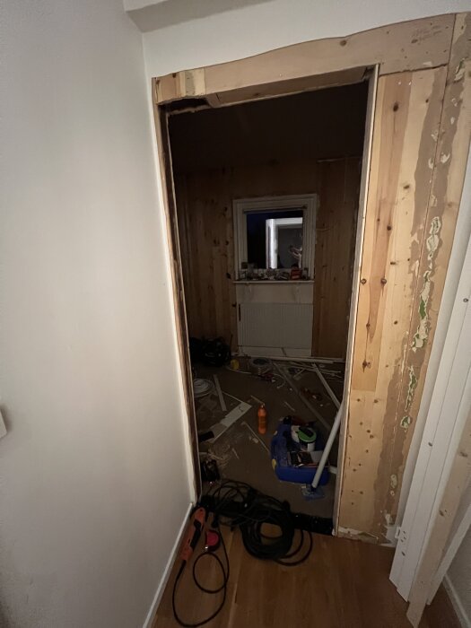 Ett dörrhål under renovering med blottade träreglar, verktyg och material på golvet, samt en rumsvy i bakgrunden.