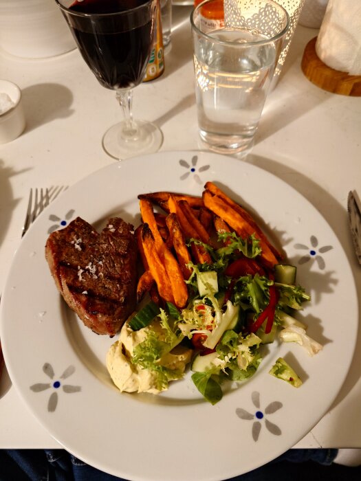 Grillad köttbit med sötpotatis-pommes, bea-sås och sallad på tallrik, rödvins- och vattenglas på bord.
