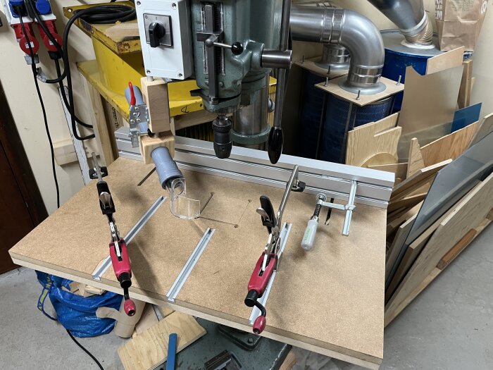 Arbetsstation med pelarborrmaskin, bord i MDF med T-spår och klämmor, samt dammhantering. Trä- och metallbearbetningsutrustning synlig i bakgrunden.