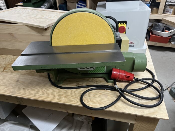 Grön och gul skivputs med röd elkabel på en träbänk i verkstadsmiljö, märkt Ejca.