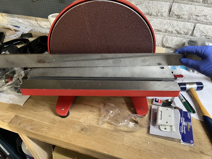 En röd skivputsmaskin med en plan yta kontrolleras med en metallrätskiva för att mäta planheten av bordet. Verktyg syns i bakgrunden.