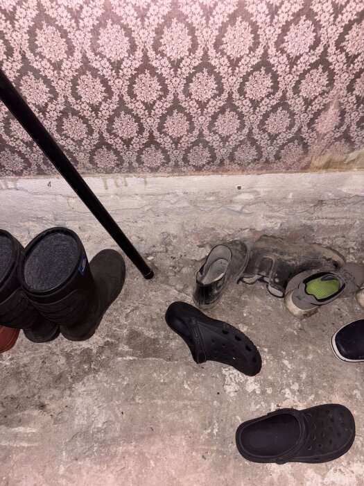 Skor och stövlar på ett ojämnt cementgolv med tapet i bakgrunden.