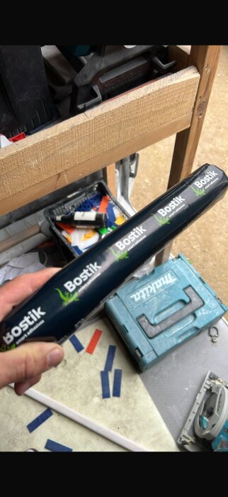 Hand håller en förpackning Bostik-lim i en verkstadsmiljö med verktygslåda och såg på arbetsbänk.