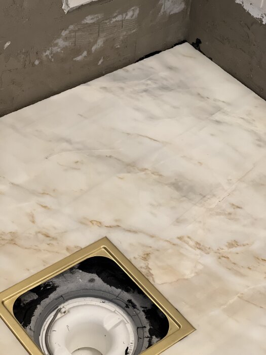 Golv i badrum med marmorliknande plattor och golvbrunn med guldram, svart tätning runt brunnen. Väggarna har spår av byggmaterial nära golvet.