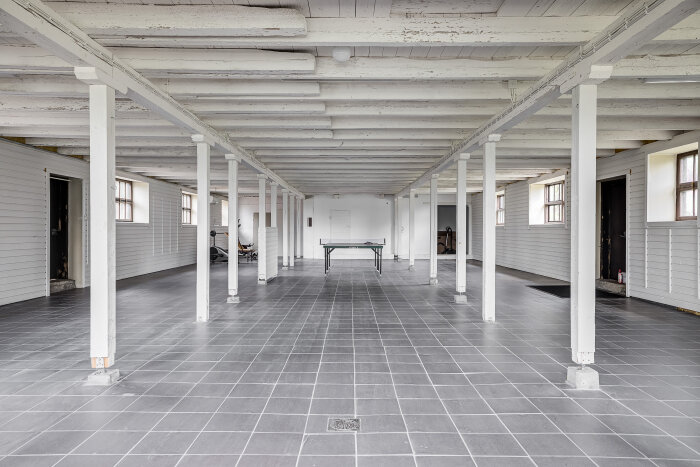 Rymlig, tom ladugård med vita pelare och grå kakelplattor på golvet. Ett pingisbord syns i mitten av rummet.