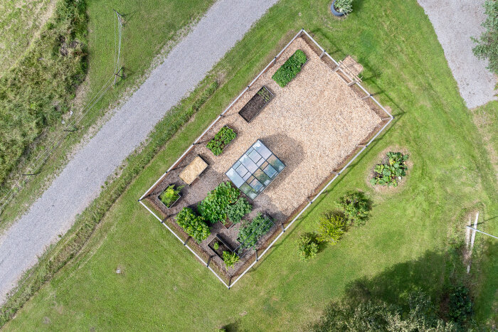 Flygbild över trädgård med inhägnad odling och växthus, omgiven av grönska och grusvägar.