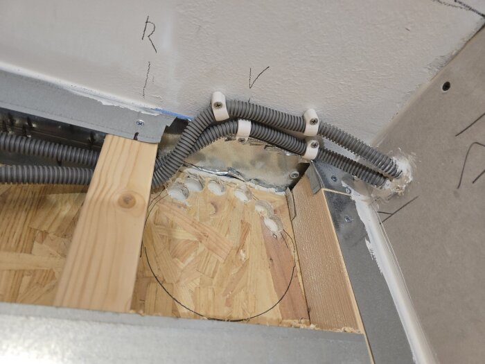 Flexrör monterade genom hål i OSB-skiva och gipsvägg för spirorör, tillsammans med träregel och metallfäste i ett byggprojekt.