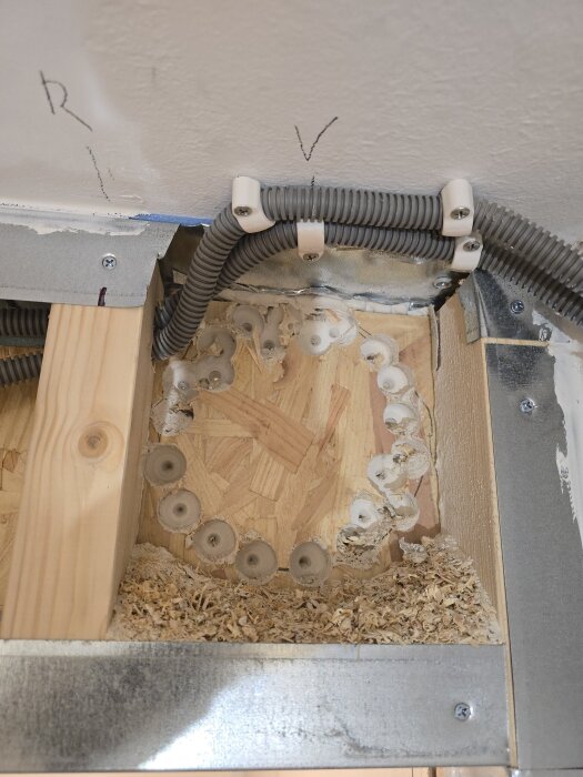 Hål borrade i OSB för spirorör, flexrör dragna ovanpå, träregel och sågspån synliga, vägg byggs med isolering och gips.