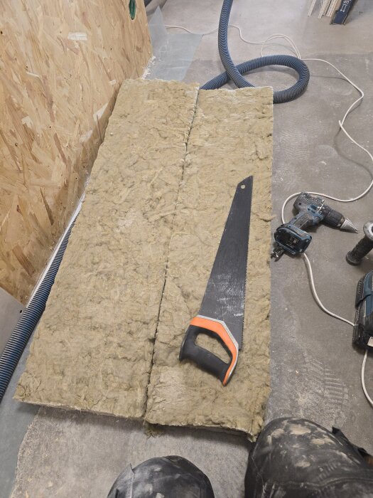 Isolering ligger på golvet med en såg ovanpå, intill en vägg av OSB-skiva. Bredvid ligger verktyg och flexrör.