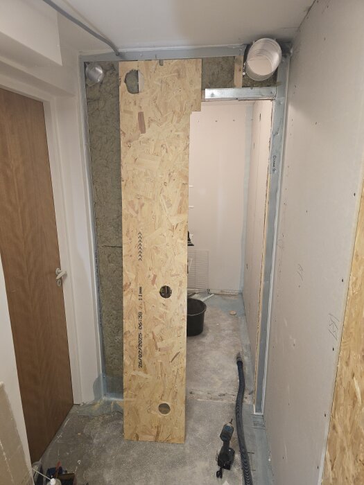 OSB-skiva med borrade hål för ventilationsrör, flexrör på golvet, väggar med isolering och gips, och en byggarbetsplats under renovering.