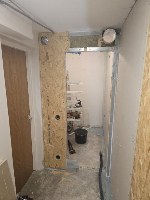 Vindhåll av trä och gips i hall, hål för spirorör borrat och flexrör draget. Ventilationskanal synlig, byggmaterial och verktyg på golvet.