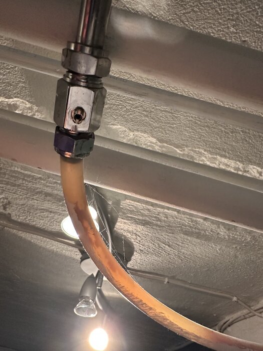 Koppling med kulventil och saknat vred under ett rör i taket, med synlig belysning i bakgrunden.