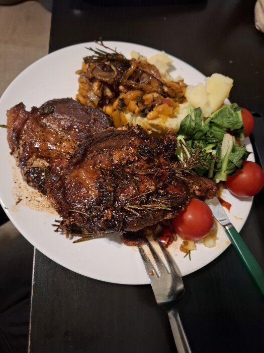 Tallrik med kött, potatismos, grönsaker och tomater, serverad med en gaffel och kniv.