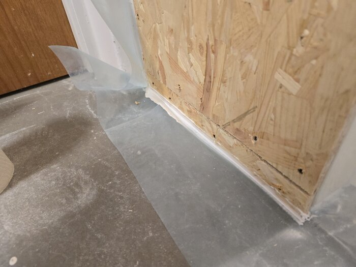 Byggplast och OSB-skiva monterade längs golvet i ett renoveringsprojekt, redo för isolering och tätning av vägg mot golv.