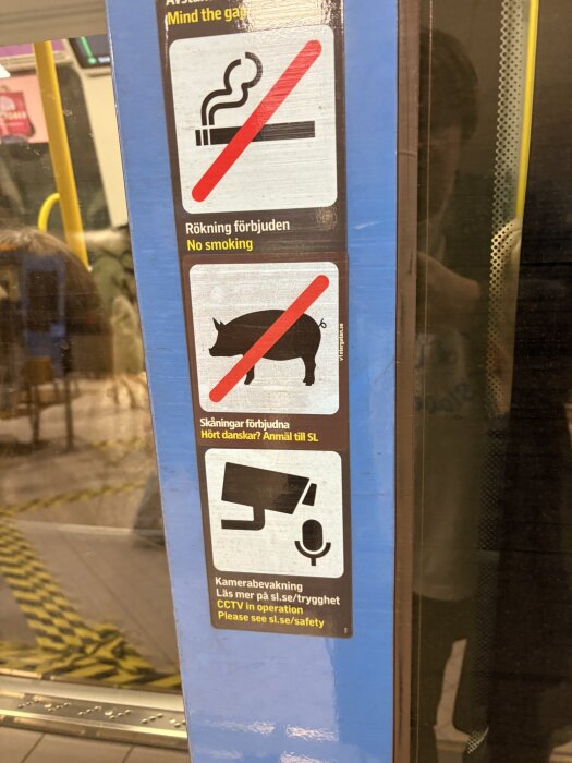 Skylt i tåg med rökning förbjuden, skämt om skåningar med överstruket gris, information om övervakning samt hänvisning till säkerhetsinformation.