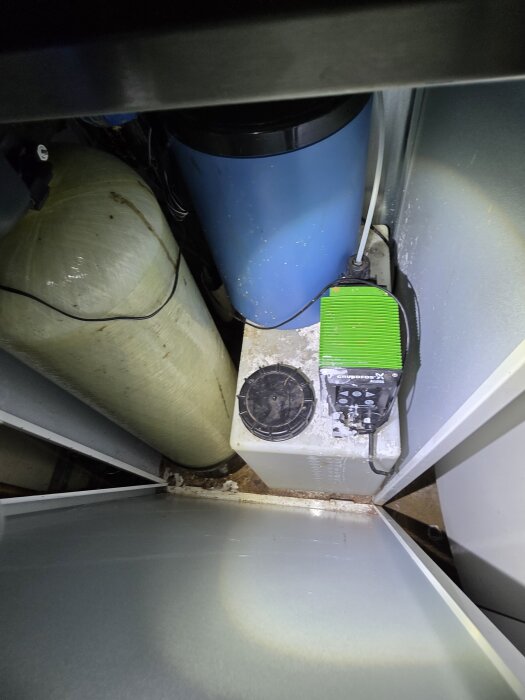 Vattenreningssystem med beige tank och blå behållare. Enhet benämnd "Erie Model 1035" syns. Gulaktiga avlagringar på golvet runt anläggningen.