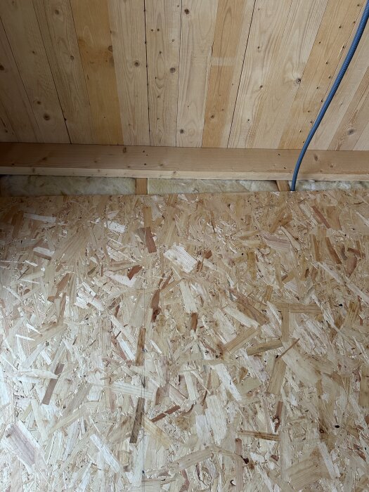 Springa mellan trägolv och trävägg, delvis fylld med isolering, med blått rör synligt på högra sidan.