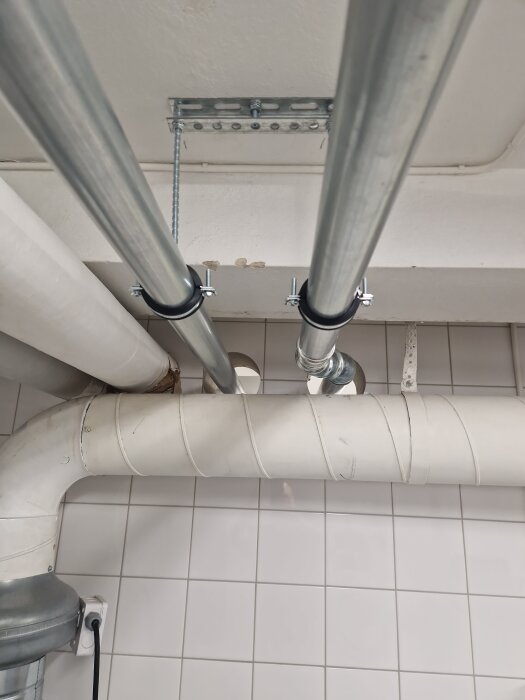 Två metallrör installerade i taket, ett rakt och ett med böjning, fastsatta med klämmor, före en kaklad vägg.