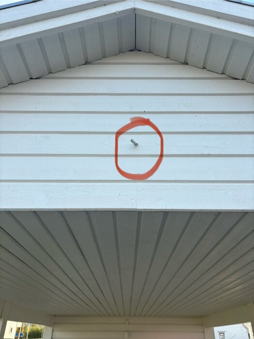 Vägg av vitmålad träpanel på carport med en elledning markerad med röd cirkel, tänkt för en övervakningskamera.