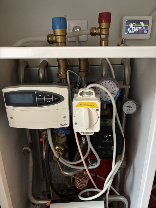 Kontrollpanel för fjärrvärmesystem med röd och blå skruvknapp, tryckmätare och kopplingar synliga.