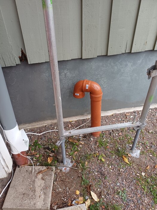 Orange markrör med 90-gradersböjning intill husvägg. Ett grått rör och en byggställning syns bredvid. Trädgårdsmaterial och stenar på marken.